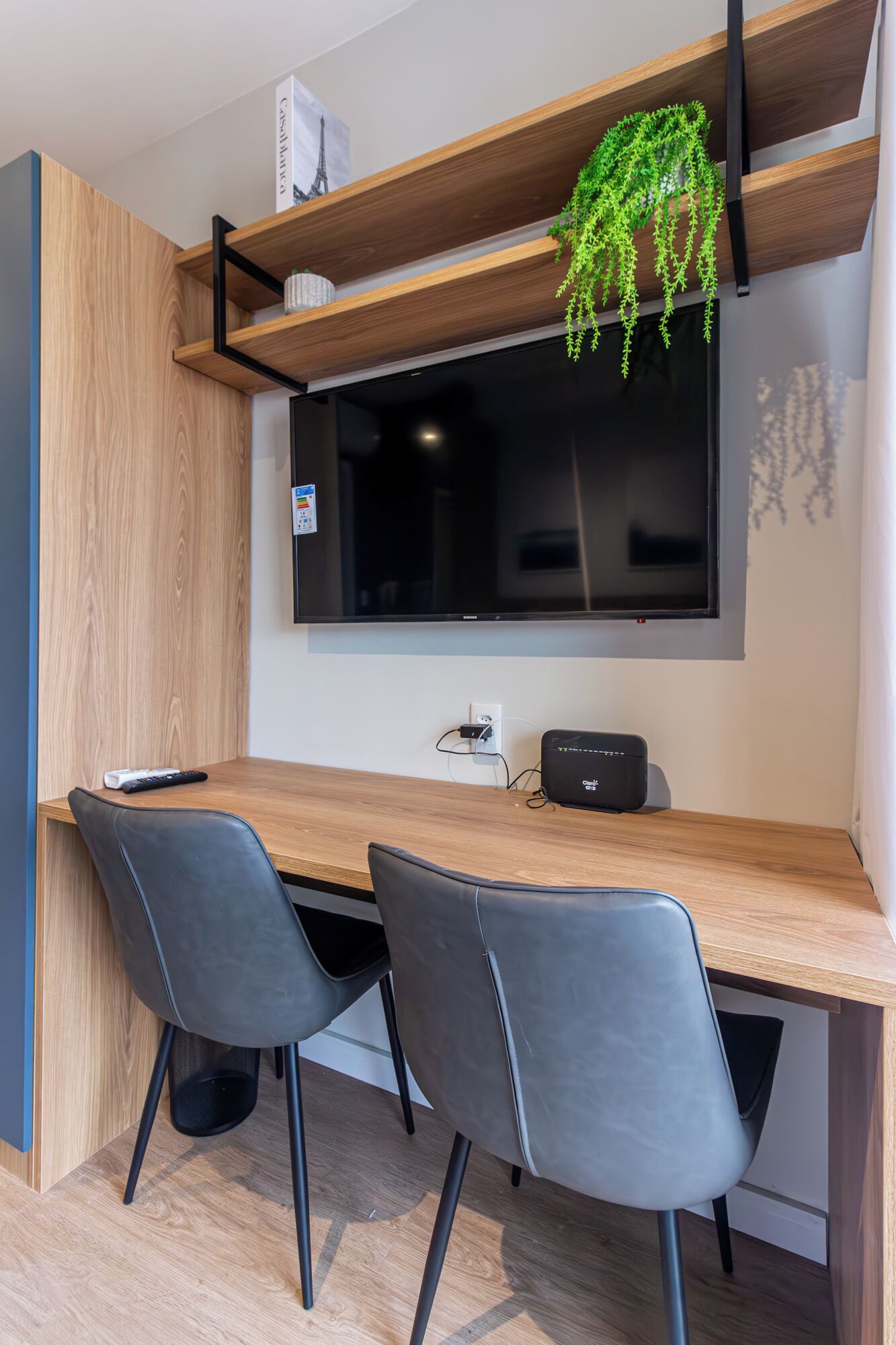 A modern dining area with a wooden table, two grey chairs, a black speaker, and a hanging plant.