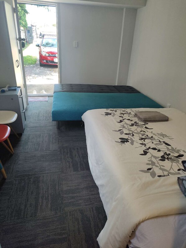 Habitación de hotel con cama, escritorio y silla. Se ve una ventana con vista a la calle y un coche estacionado afuera.