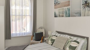 Een slaapkamer met een bed, een stoel, een raam met gordijnen en wanddecoratie met strandtaferelen.