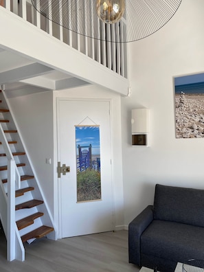 Un escalier avec des marches en bois et des murs blancs, une porte ornée d’une affiche, un canapé gris et un luminaire au plafond.