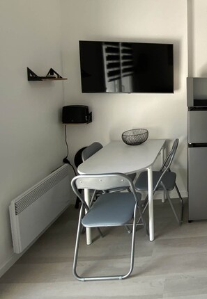 Un comedor moderno con una mesa blanca, dos sillas plegables, un altavoz negro, un televisor montado en la pared y un estante con un adorno.