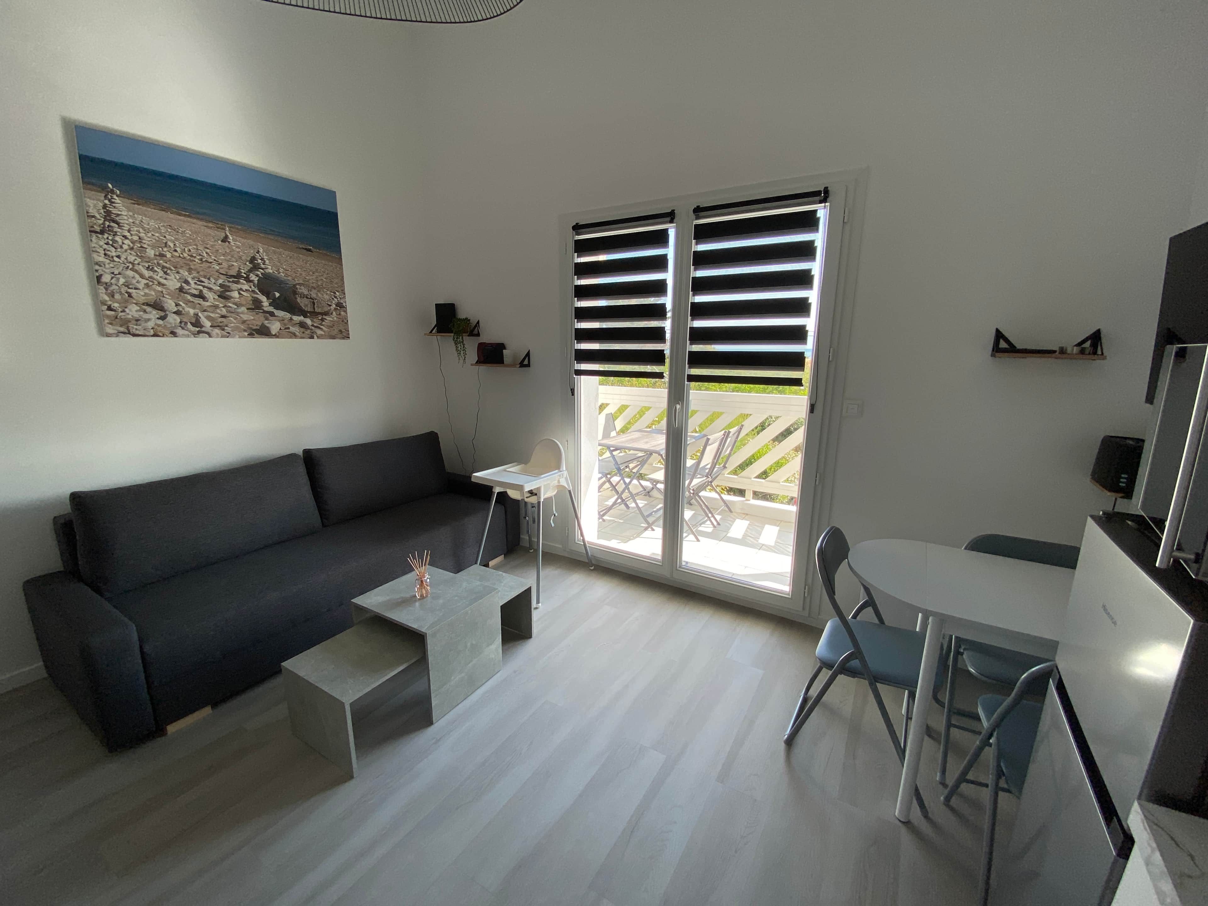 Ein modernes Wohnzimmer mit einer Couch, einem kleinen Tisch, einem Sessel, einem Fernseher und einer Glastür zum Außenbereich.