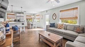 Un vaste espace de vie comprenant un canapé, une table basse et une table à manger. On y trouve également un ventilateur de plafond, des luminaires suspendus et de grandes fenêtres équipées de stores.