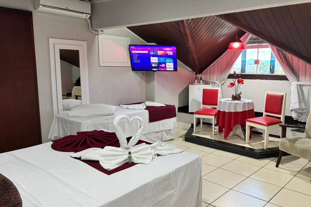 Ein Hotelzimmer mit zwei Betten, einem Fernseher, einer kleinen Essecke mit roten Stühlen und Blick durch ein Fenster.