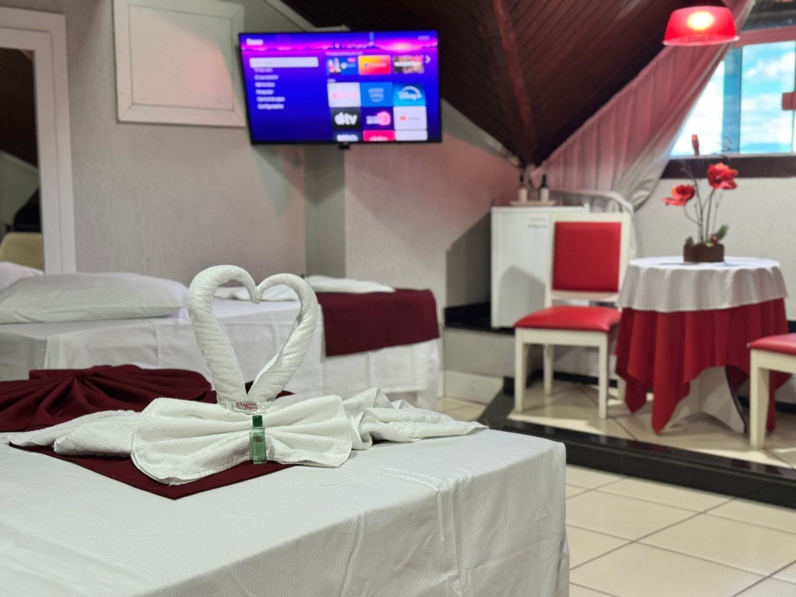 Una habitación de hotel con una cama, un televisor montado en la pared, una mesita con mantel rojo y una silla.