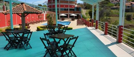 A covered outdoor area with tables and chairs, a pool, and a view of a red building in the background.