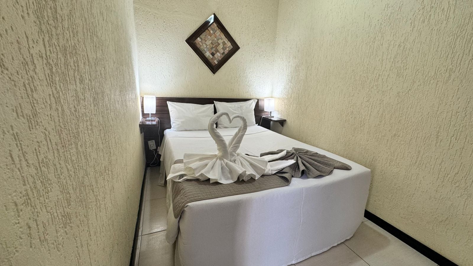 A hotel room with a neatly made bed, a bedside table with a lamp, and a wall-mounted picture frame.