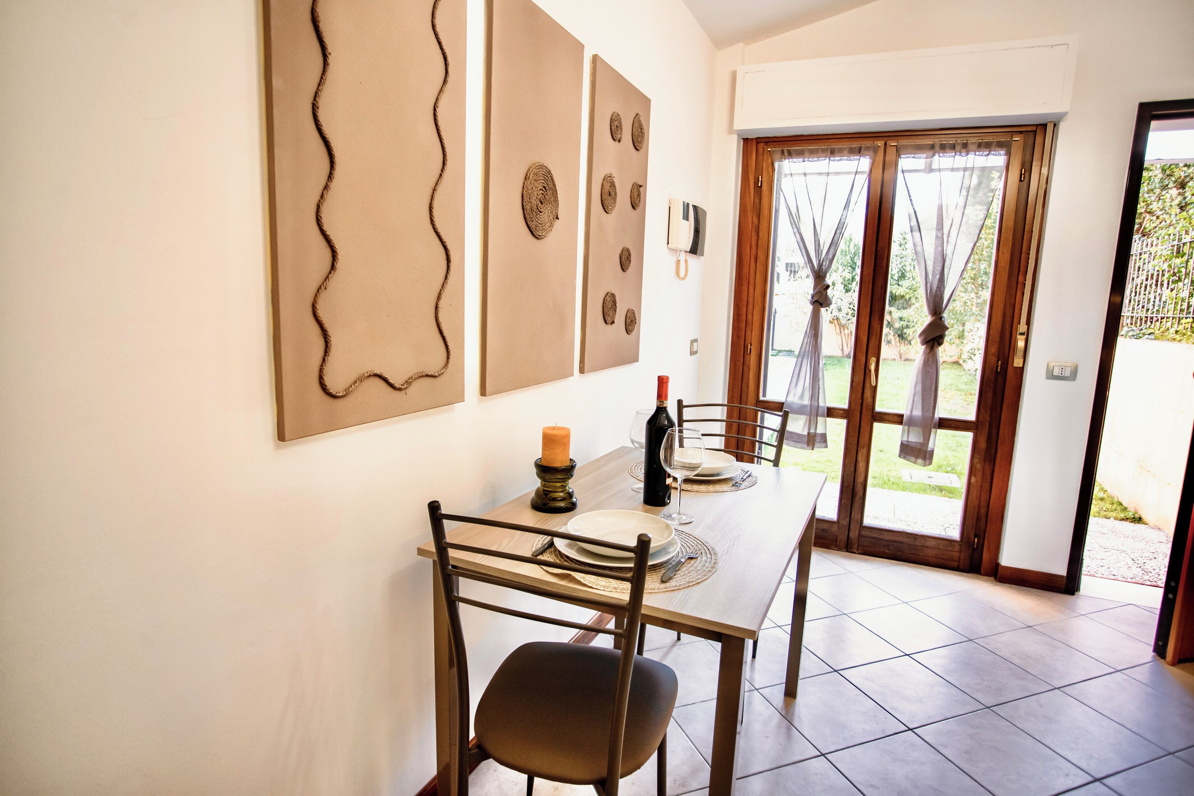 Een eethoek met een tafel gedekt voor twee personen, een kaars en een fles wijn. Er staan twee stoelen bij, een wandlamp en een deur naar buiten.