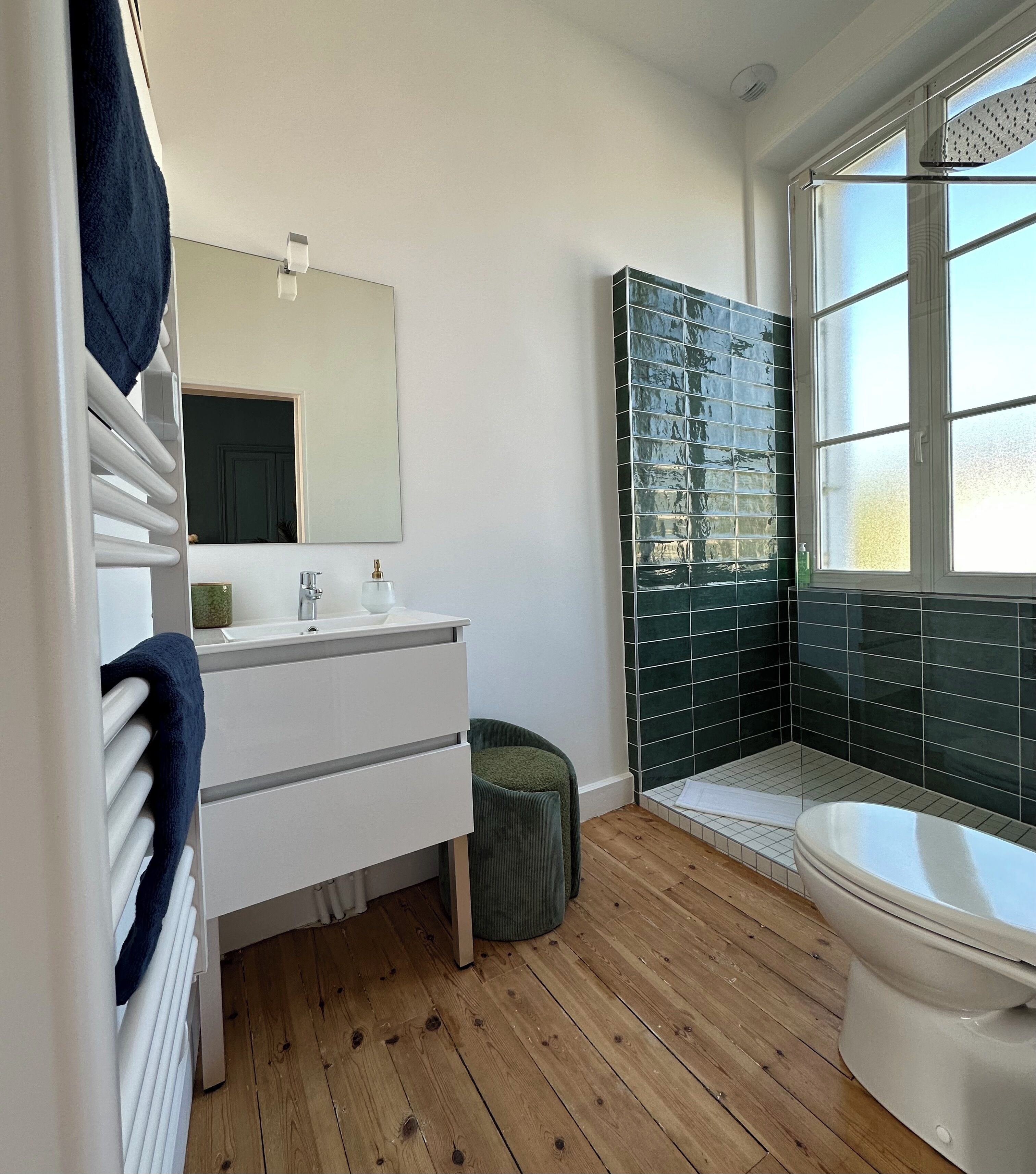 Een moderne badkamer met een wit wastafelmeubel, een grote spiegel, een douchegedeelte betegeld met groene tegels en een raam voor natuurlijk licht.