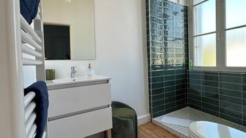 Een moderne badkamer met een wit wastafelmeubel, een grote spiegel, een douchegedeelte betegeld met groene tegels en een raam voor natuurlijk licht.