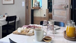 Ein Essbereich mit einem Tisch, der für ein Frühstück gedeckt ist, einschließlich Käse, Fleisch und Brot. Es gibt Tassen, eine Kanne Orangensaft und eine Kaffeemaschine. Der Raum hat eine Holztür, ein gerahmtes Bild an der Wand und einen Blick in einen anderen Raum mit einem blauen Vorhang.