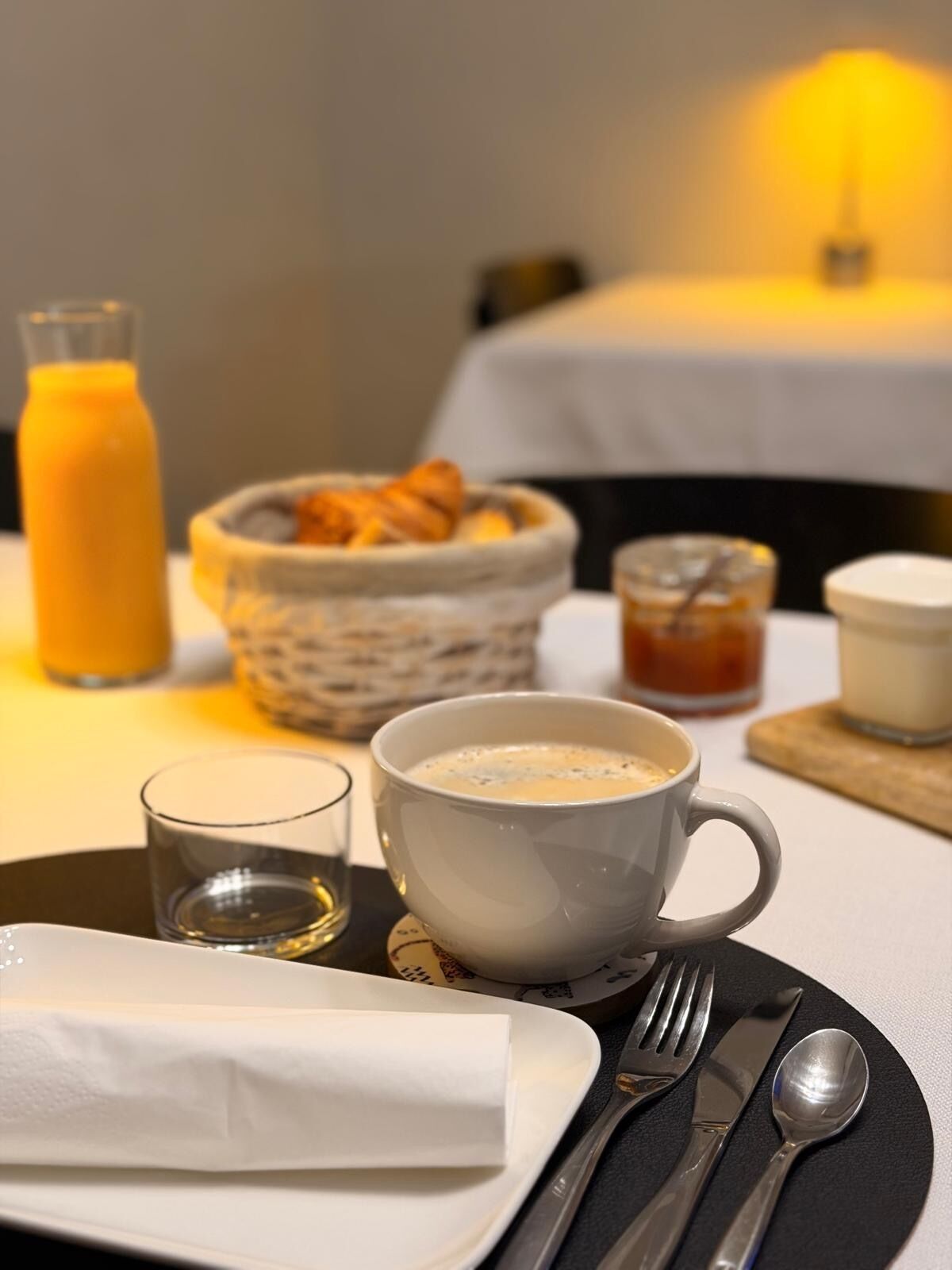 A breakfast setting with a cup of coffee, a glass of orange juice, a bowl of croissants, and a small jar of jam on a table.