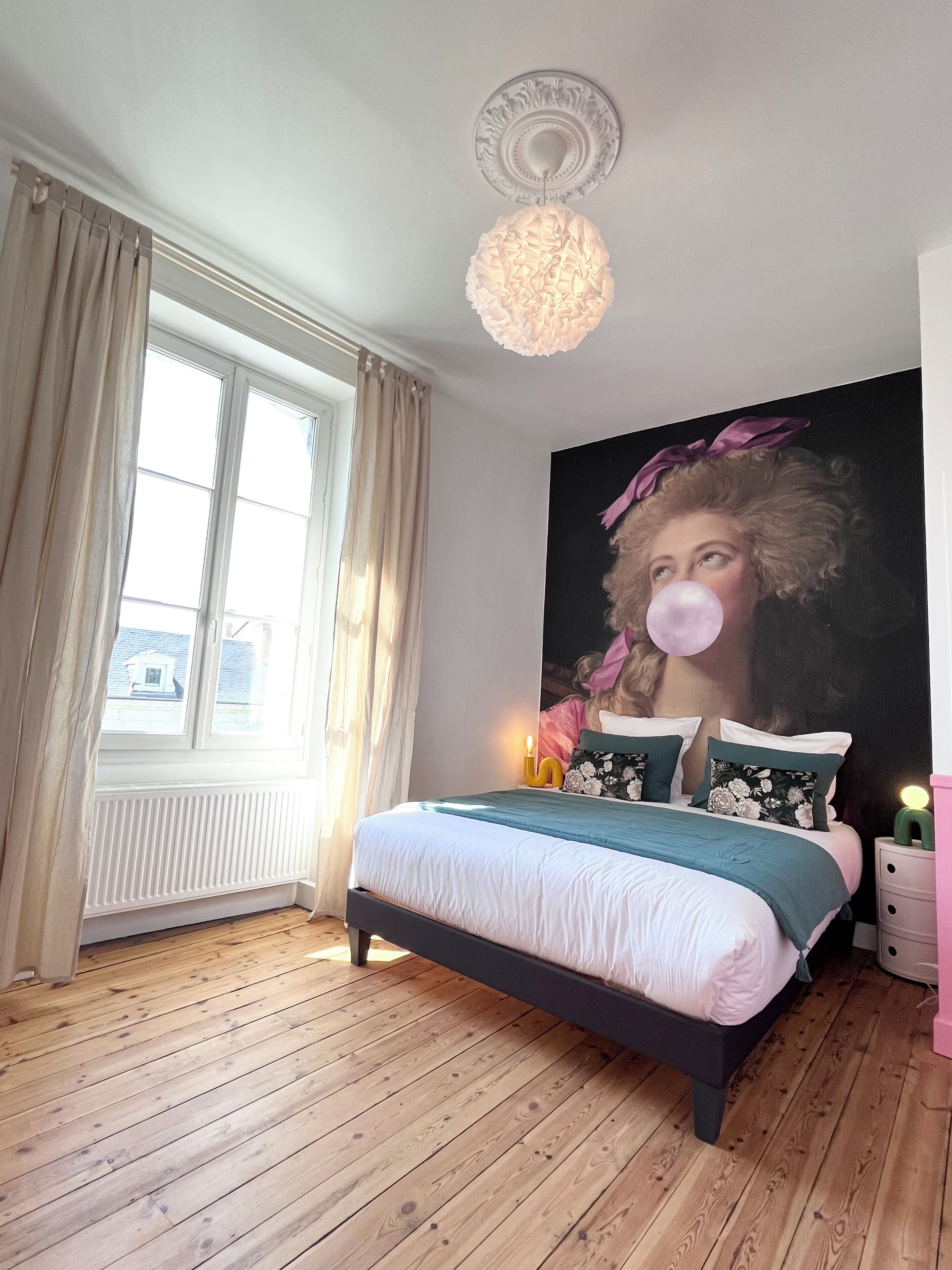 A bedroom with a large bed, a window with curtains, a wall-mounted light fixture, and a painting.
