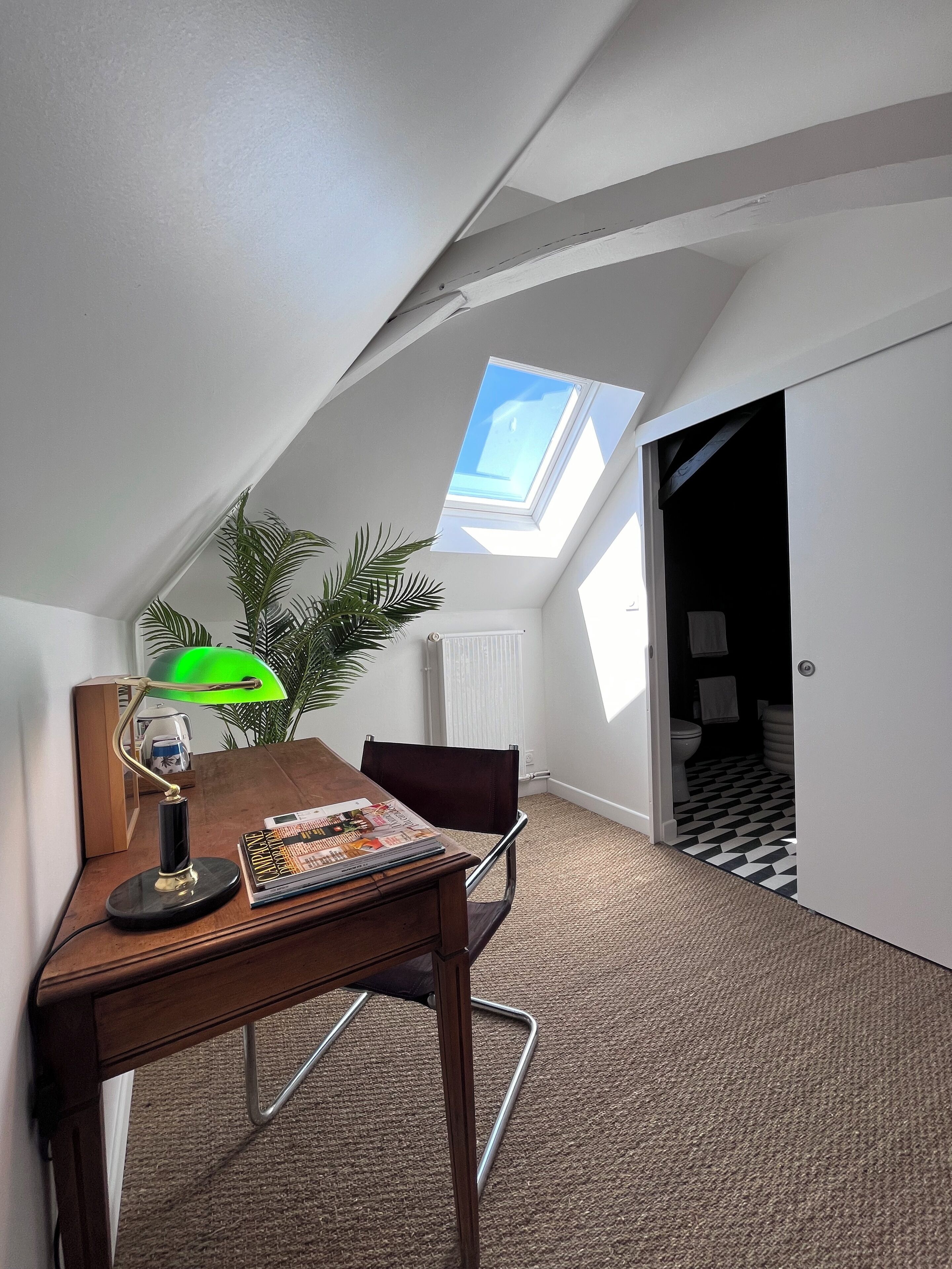 Una habitación con techo inclinado, tragaluz, escritorio con lámpara, silla y planta en maceta.