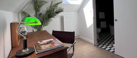 Una habitación con techo inclinado, tragaluz, escritorio con lámpara, silla y planta en maceta.