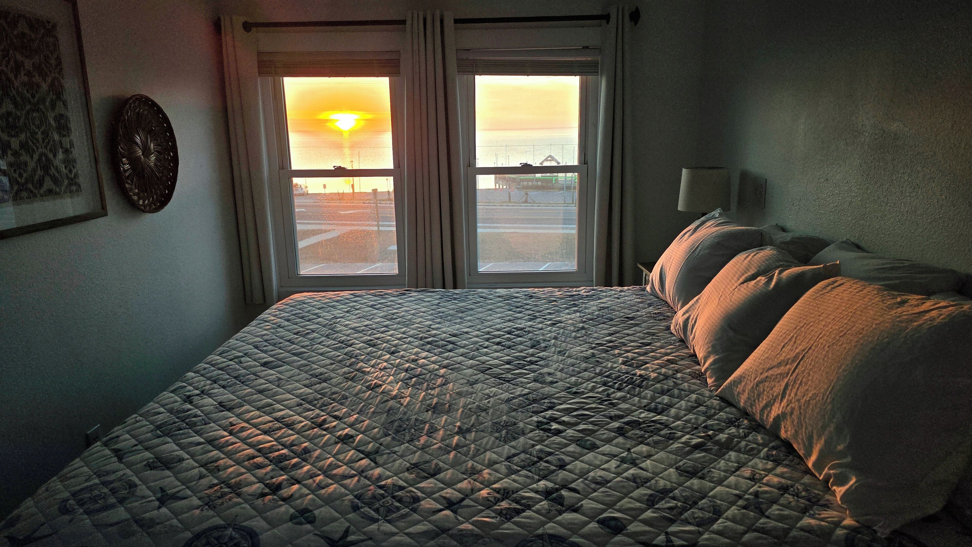A bedroom with a bed, a window with a view of the sunset, and a wall-mounted light fixture.