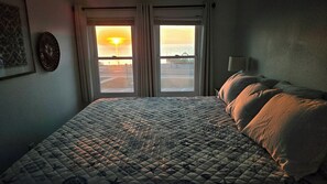 A bedroom with a bed, a window with a view of the sunset, and a wall-mounted light fixture. - Steps to Duck Fun: Sunset Views, Resort Amenities! w/Beach Access! (Duck)