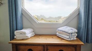 Une commode en bois surmontée de deux piles de serviettes blanches, une fenêtre donnant sur des arbres et des rideaux bleus.