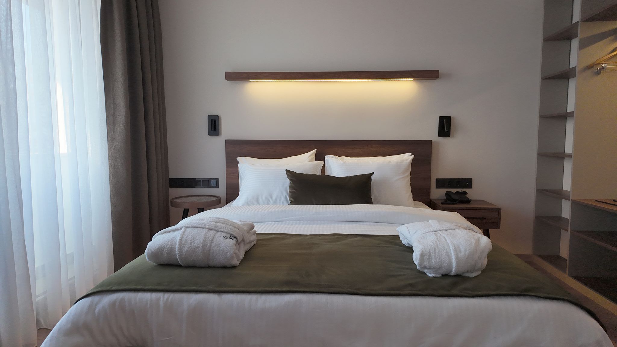 A hotel room with a large bed, a bedside table, a nightstand, a wall-mounted light, and a closet with shelves.
