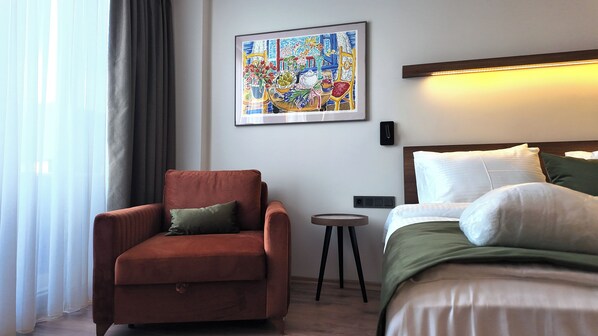 A hotel room with a bed, a chair, a small table, and a painting on the wall.