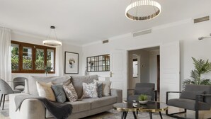 Ein modernes Wohnzimmer mit einem grauen Sofa, einem runden Couchtisch und einem grauen Sessel. Zwei Pendelleuchten befinden sich über dem Sofa und ein großes Fenster mit Vorhängen.