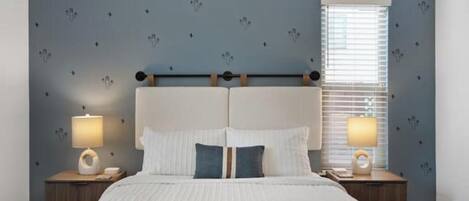 A bedroom with a bed, two bedside tables with lamps, a window with blinds, and a wall with a cactus pattern.