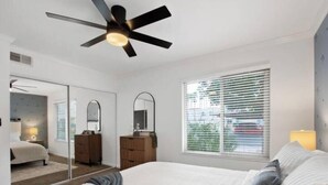 Ein Schlafzimmer mit einem Bett, einem Deckenventilator, einem Schminktisch, einem Spiegel und einem Fenster mit Jalousien.