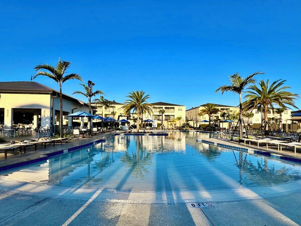 A swimming pool with lounge chairs and umbrellas, surrounded by palm trees and hotel buildings.