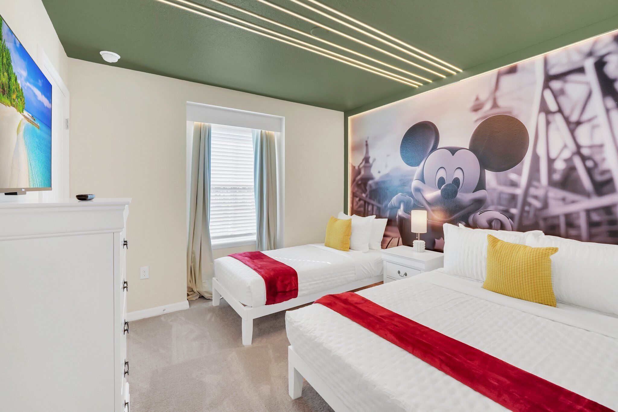 Ein Hotelzimmer mit zwei Betten, einem großen Wandbild mit Mickey Mouse, einem Fernseher und einem Fenster mit Vorhängen.