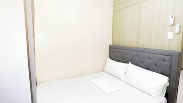Cama bem arrumada com lençóis brancos num quarto com paredes claras e um detalhe em painel de madeira.