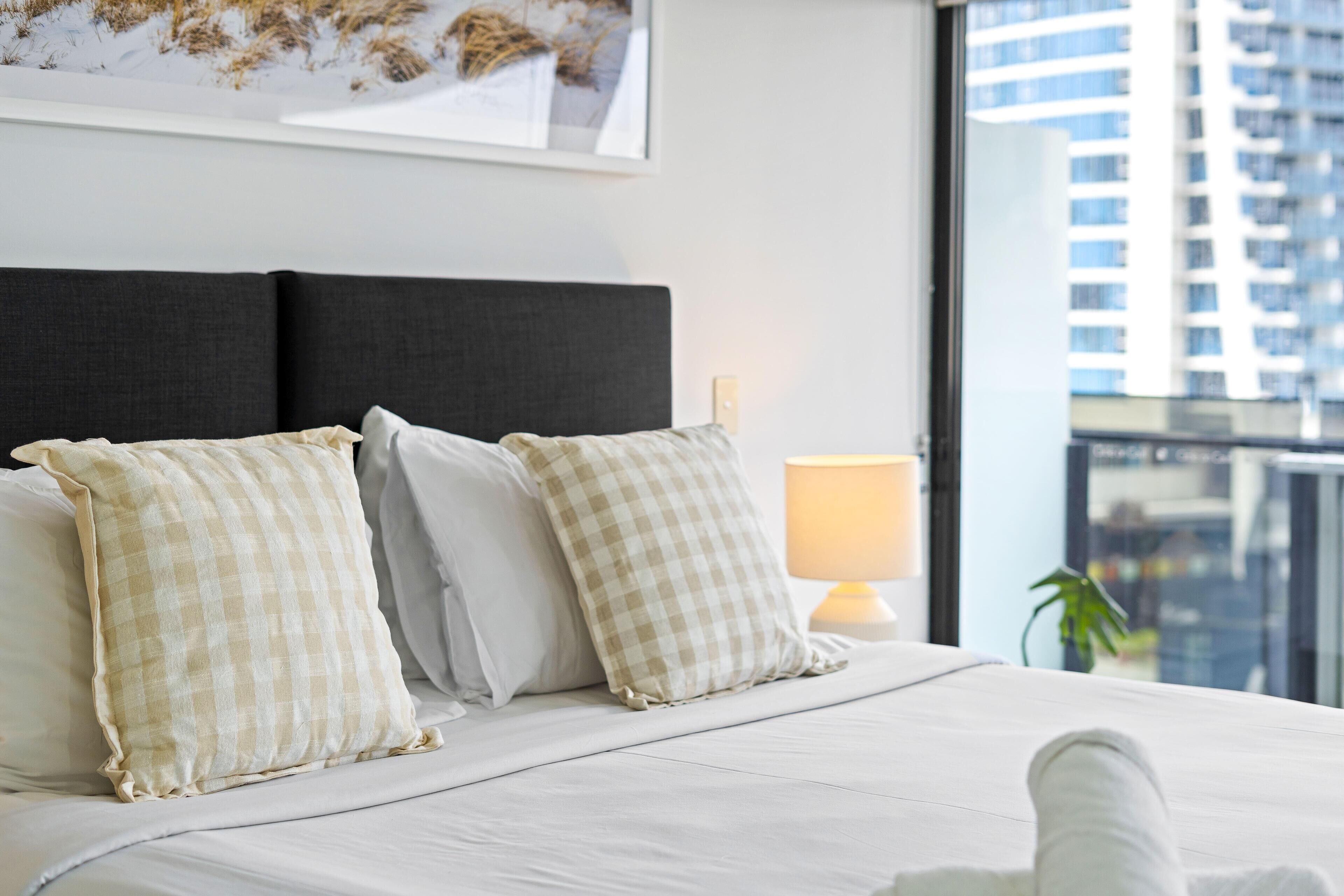 A hotel room with a large bed, checkered pillows, a bedside lamp, and a view of a cityscape through a large window.