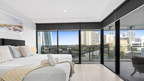 A modern bedroom with a large bed, a city view through floor-to-ceiling windows, and a balcony with a wicker chair.