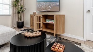 A living room with a black round coffee table, a wooden cabinet with a Monopoly board game, and a large flat-screen TV displaying a beach scene. - Summer Safari - GORGEOUS Home Near Busch Gardens & USF (Tampa)