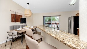 A modern kitchen with a granite countertop, a dining area with a table and chairs, and a view of the outdoors through a window. - Summer Safari - GORGEOUS Home Near Busch Gardens & USF (Tampa)