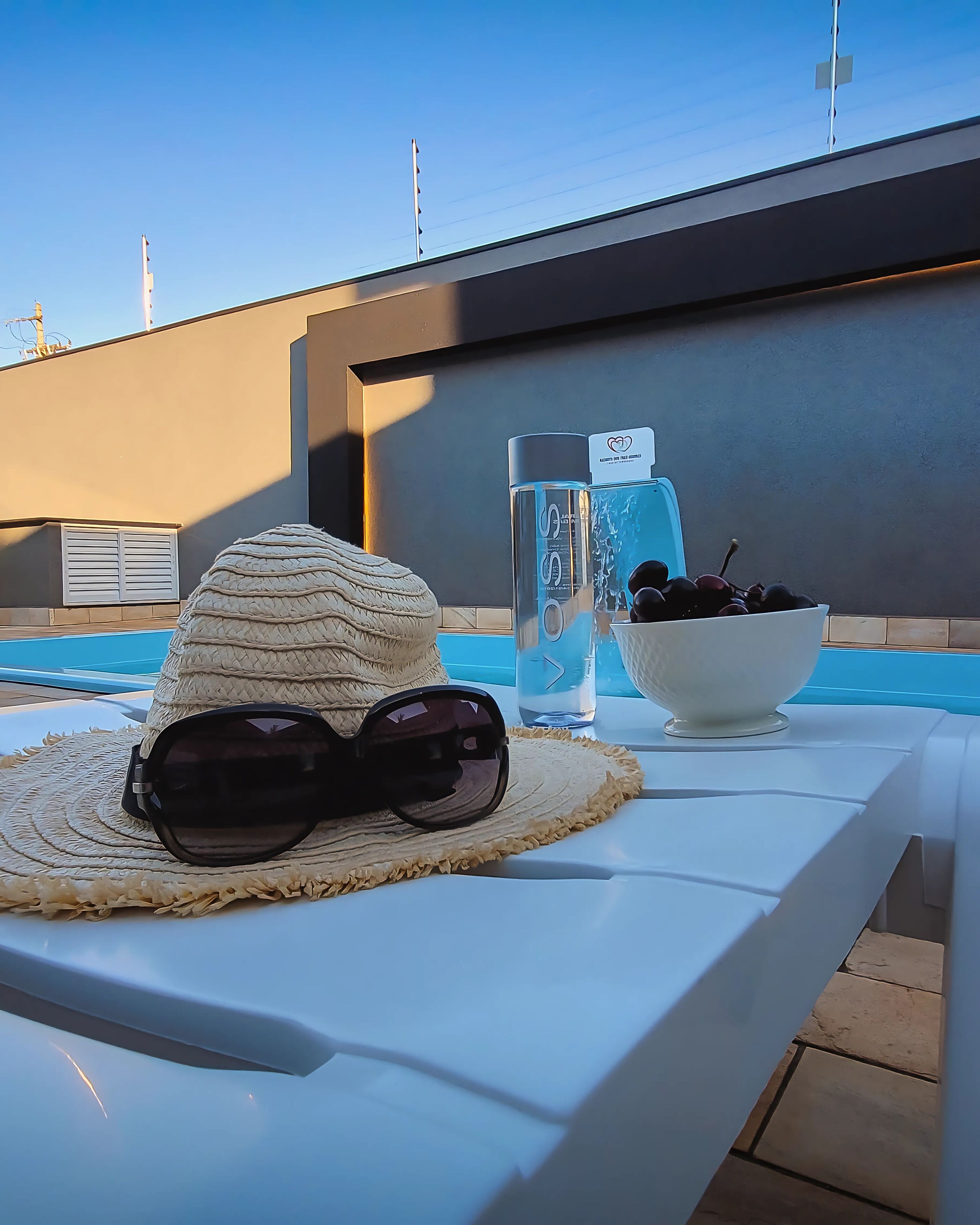 Escena a la piscina amb un barret de sol, ulleres de sol, una ampolla d'aigua i un bol de fruita damunt d'una taula blanca.