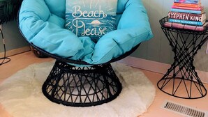 A blue cushioned chair with a pillow that reads "Beach Please" on a white furry rug.