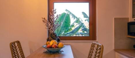 Un comedor con mesa de madera, dos sillas de mimbre, un cuenco de fruta y una ventana con vistas a follaje tropical.