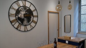 A dining area with a wooden table, clear chairs, a large wall clock, and hanging light bulbs.