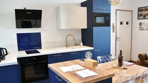 A modern kitchen with a built-in oven, microwave, and sink. There is a wooden island with a table and chairs, and a bottle of wine on the counter.