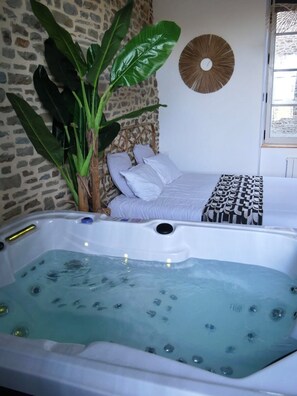 Une pièce avec un grand jacuzzi, un lit avec du linge de lit blanc, une plante en pot et une horloge murale.