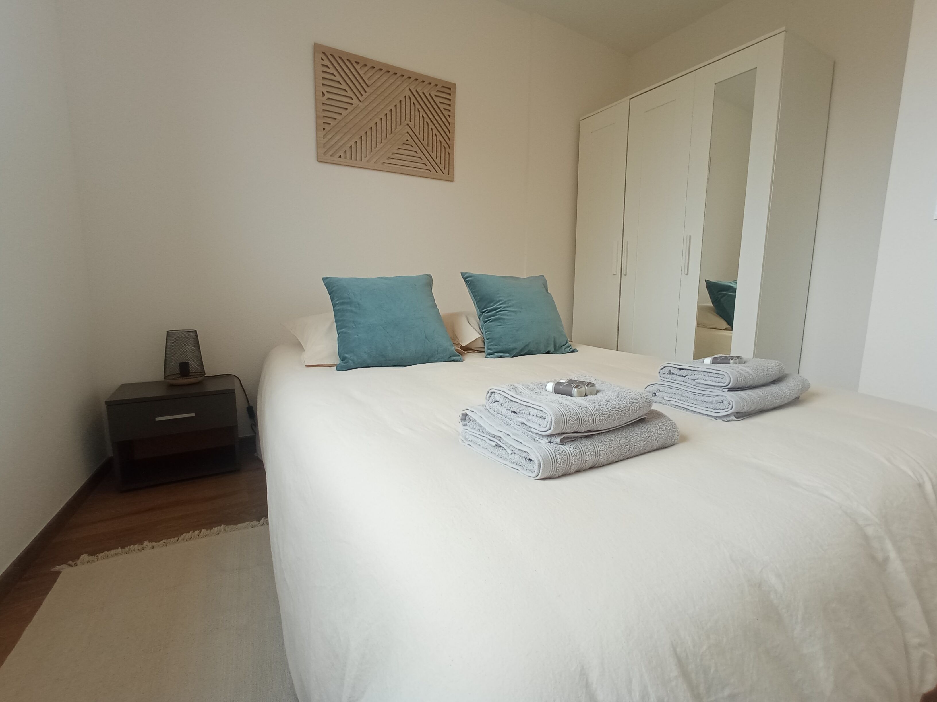Een net opgemaakt bed met witte lakens en twee blauwgroene kussens, een nachtkastje met een lamp en een garderobe met een spiegel.