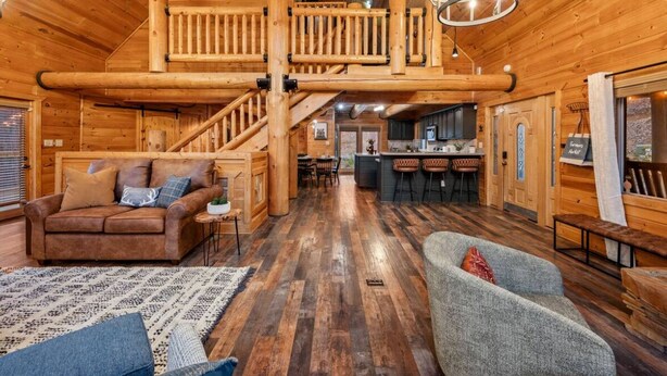 A log cabin interior with a wooden staircase, a living room with a sofa and armchair, a kitchen area, and a bar. - Large Game Garage, Fire Pit, Hot Tub, Mt. LeConte Views! (Sevierville)