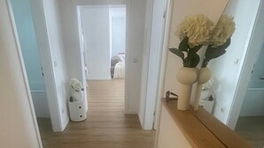 Un couloir étroit au plancher de bois mène à une chambre. De chaque côté, on trouve des portes blanches, une petite armoire blanche ornée d’un vase de fleurs et un miroir sur le mur de droite.