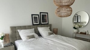 Une chambre avec un grand lit, deux tables de chevet, un luminaire suspendu en osier, un miroir rond et des tableaux encadrés accrochés au mur.