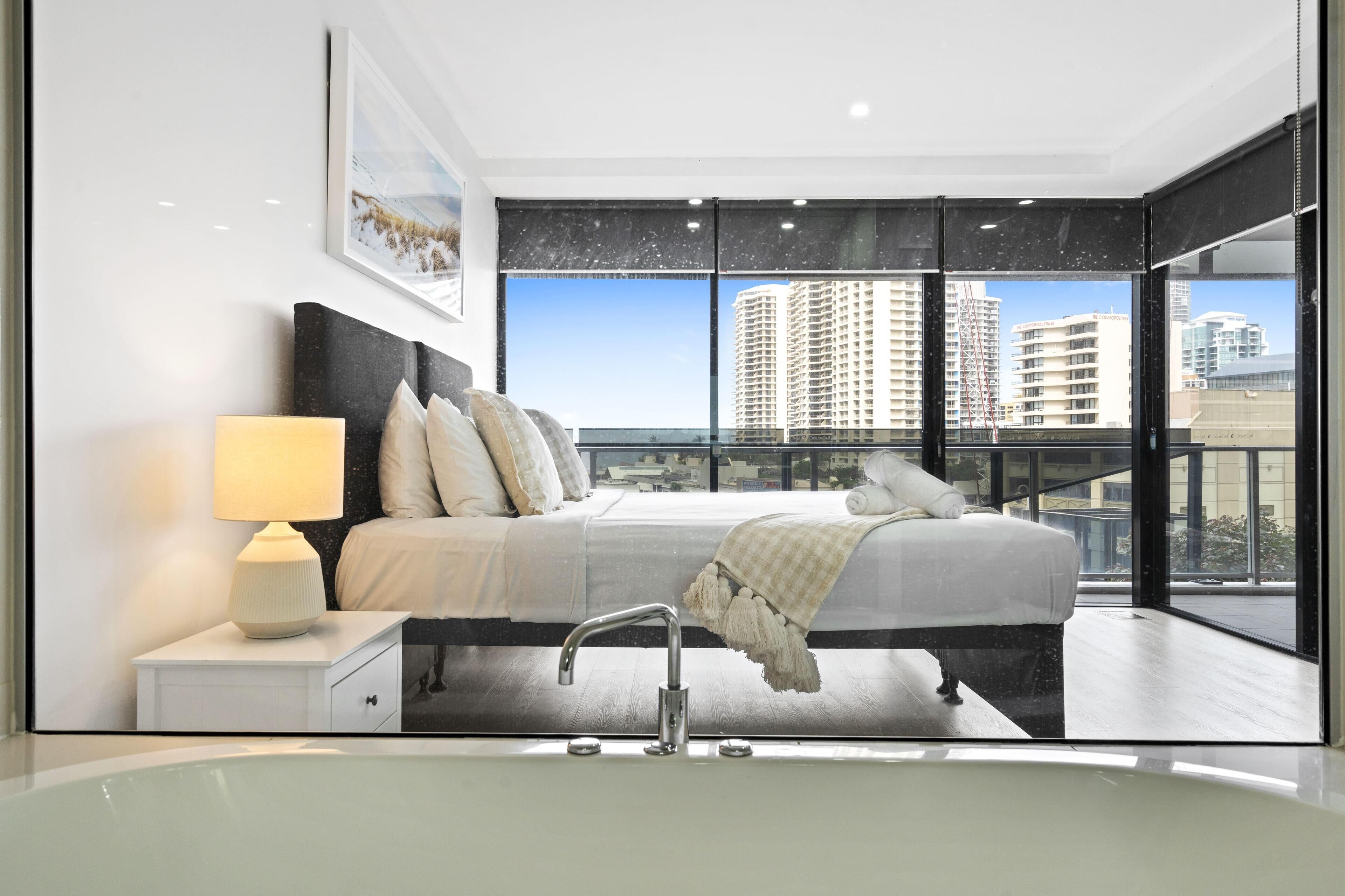 Un dormitorio moderno con una cama grande, una lámpara de noche y vistas de edificios altos a través de ventanas de gran altura.