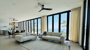 Una sala moderna con dos sofás, un comedor y grandes ventanales con vistas al mar.