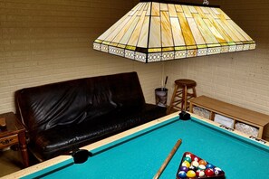 A pool table with a pool cue and balls, a leather sofa, a wooden stool, and a stained glass lamp.