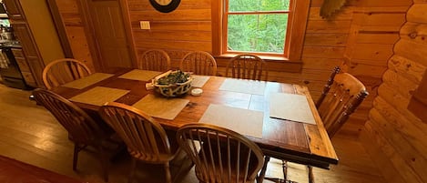 A wooden dining room with a large table, chairs, a clock, and a window with a view of trees.
