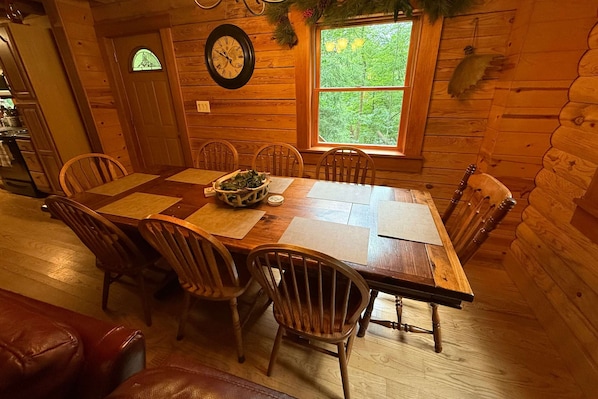 Ein Esszimmer aus Holz mit einem großen Tisch, Stühlen, einer Uhr und einem Fenster mit Blick auf Bäume.