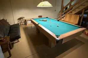 Ein Billardtisch in einem Raum mit einer Holzbank, einem kleinen Hocker und einer Pendelleuchte.